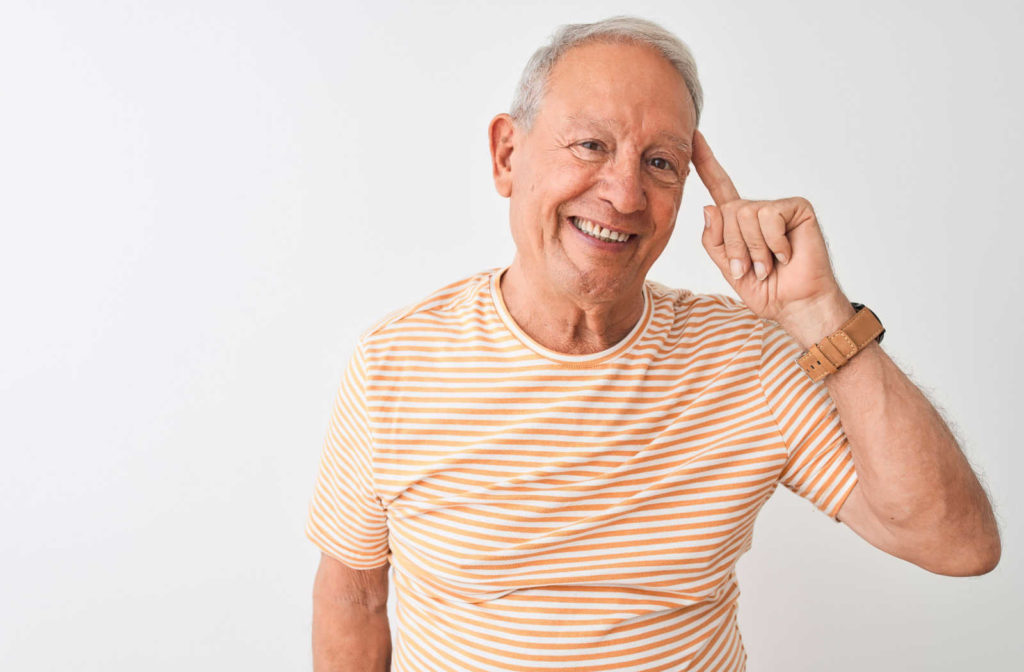 A close-up portrait of a senior man with grey hair wearing a striped t-shirt, staring into the camera with a happy expression and pointing to his head.