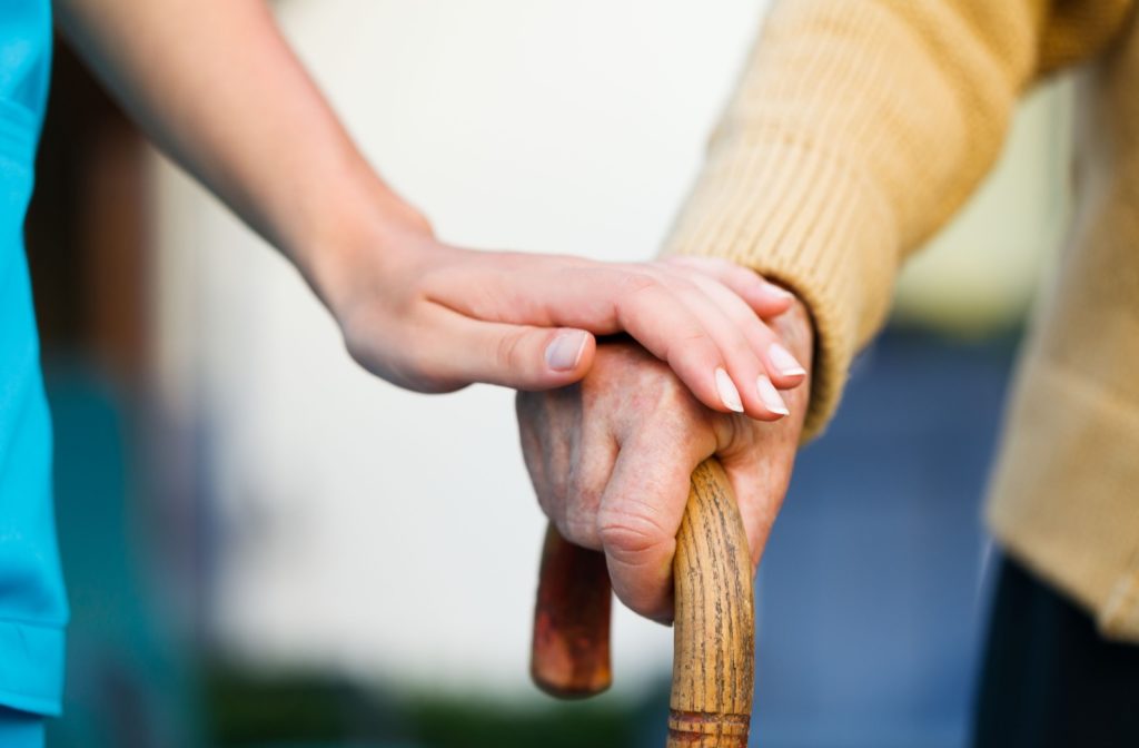 A nurse's hand on top of a senior's hand resting on a walking cane to showcase support and empathy.