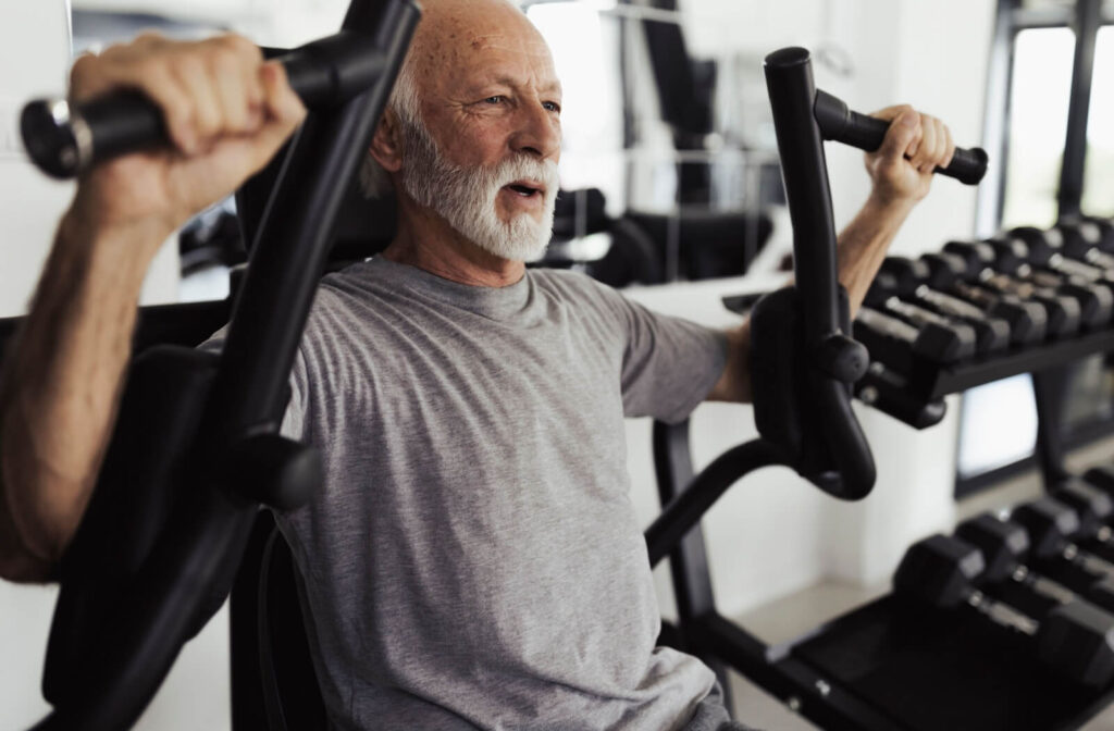 An older adult wearing a grey T-shirt uses a chest press machine in a fitness center, focusing on strength training.