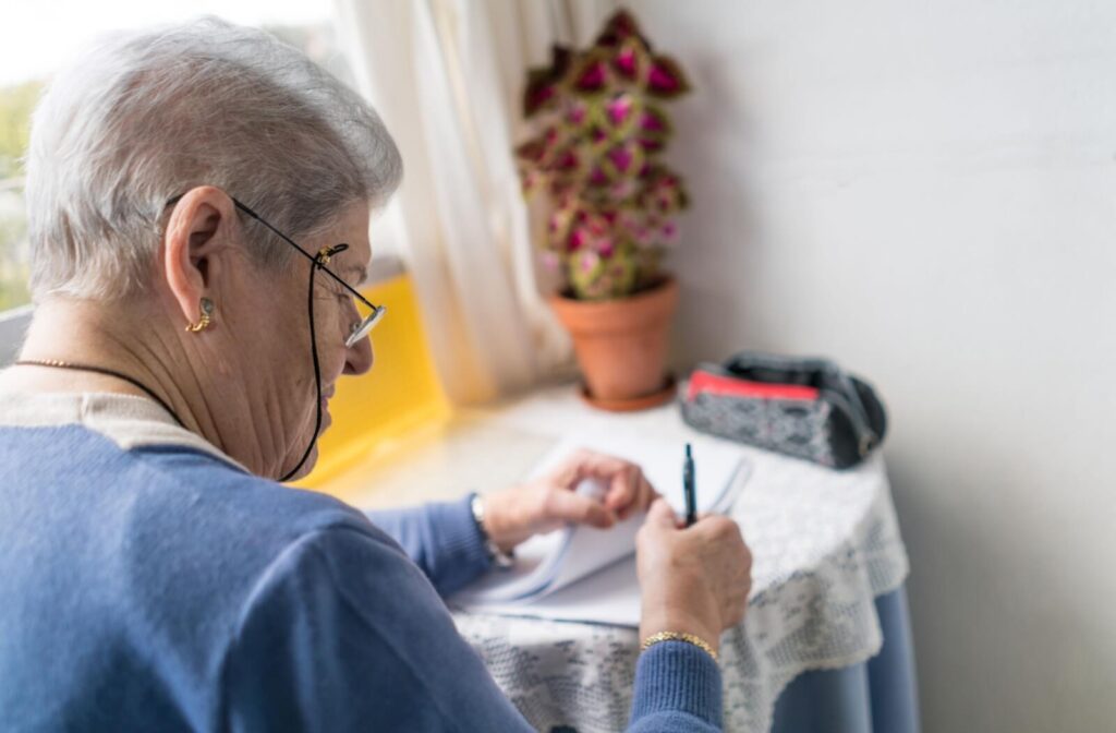 Older adult seated at a small table writing in a notebook near a window with a potted plant and glasses resting on their face