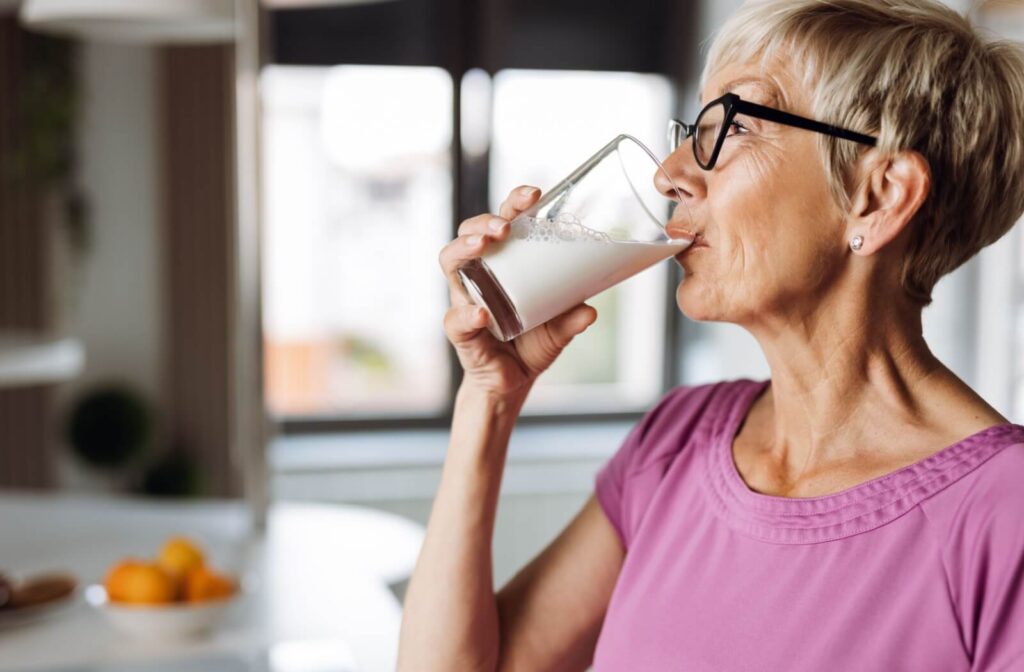 An older adult wearing glasses drinks a full glass of milk in a kitchen setting.