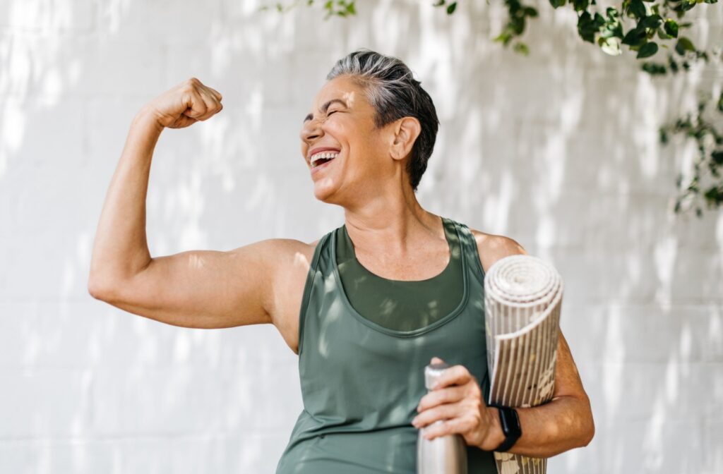 A smiling older adult woman flexes her arm muscles after a strength training session.
