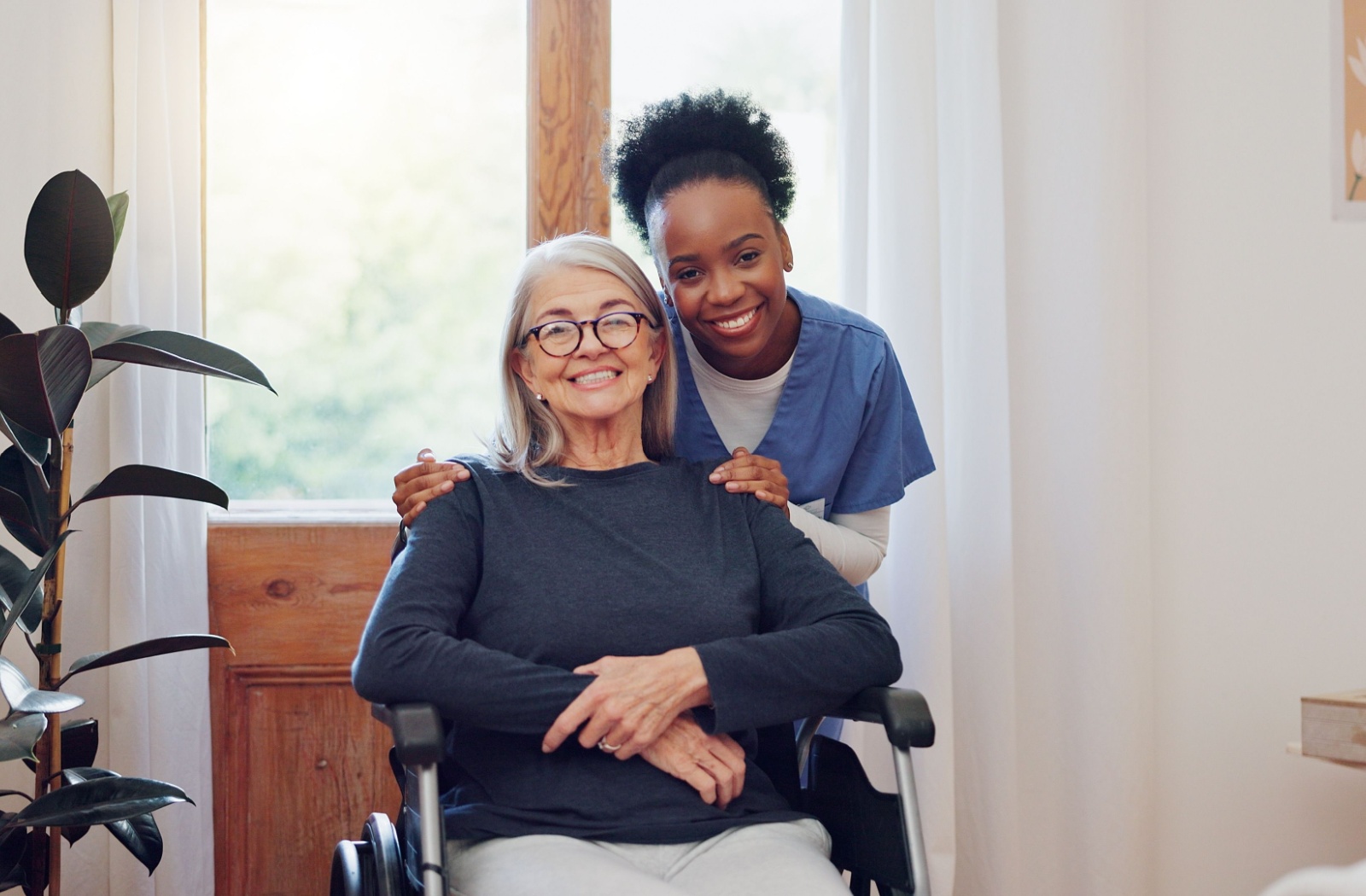 A smiling caregiver stands behind a smiling senior in a wheelchair at an assisted living community.