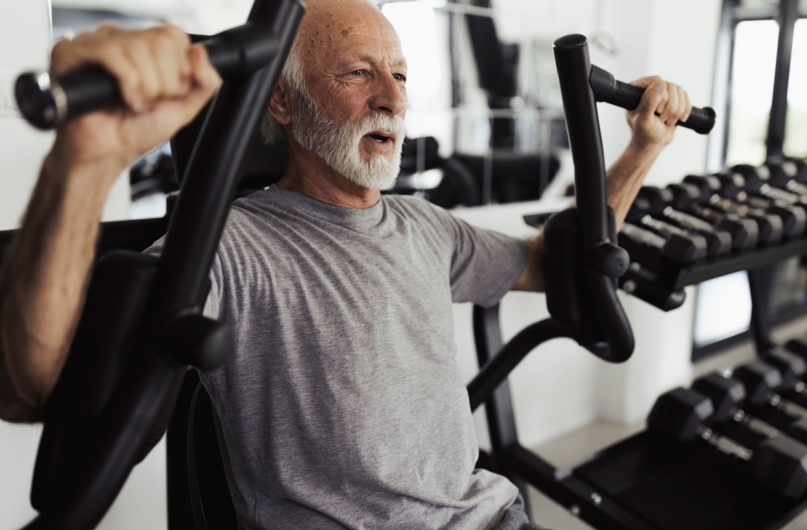 A senior uses weight-lifting equipment for strength training in a bright gym setting.