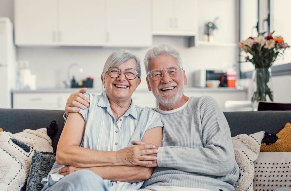 An older couple laugh and smile while sitting on a couch with arms around one another in their new living room in senior living