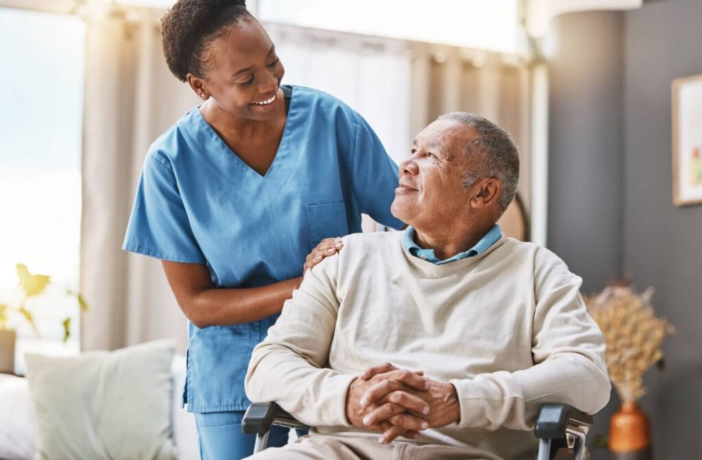 A nurse in blue scrubs smiles at a senior in a wheelchair, both happy with how well post-surgery recovery is going