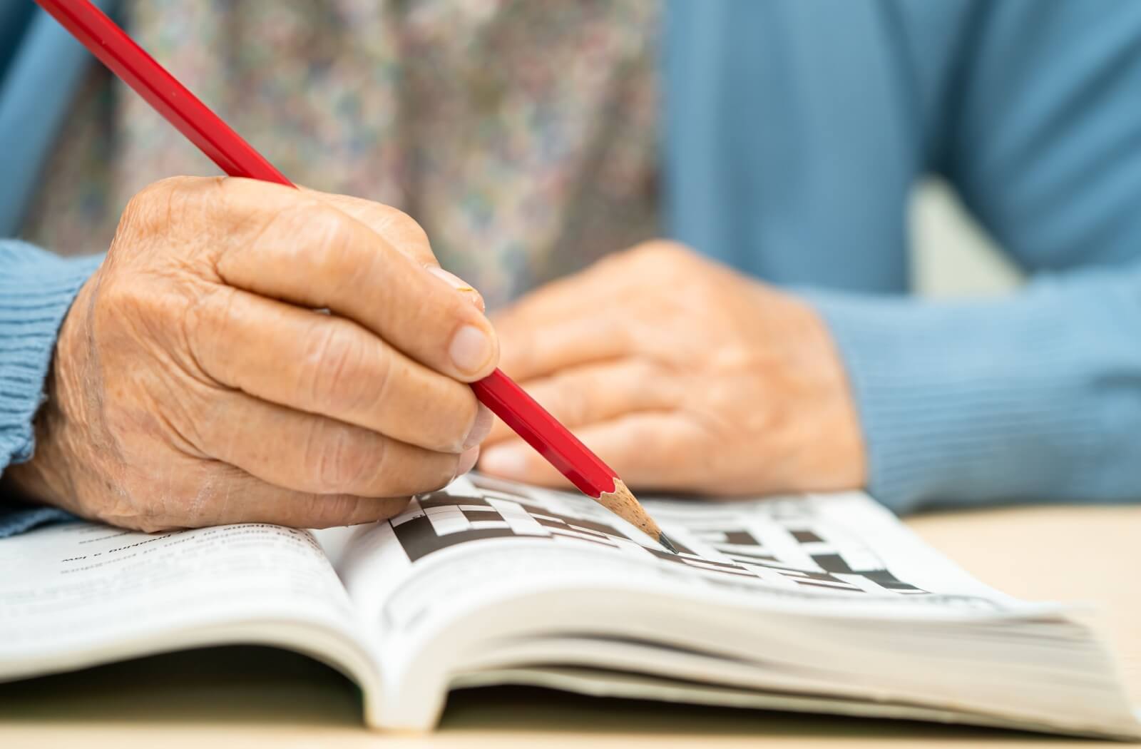 Close-up of an older adult using a red pencil to complete a physical crossword puzzle in a book